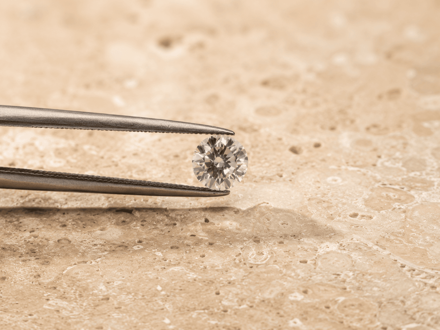 A round diamond held in tweezers on a warm stone surface